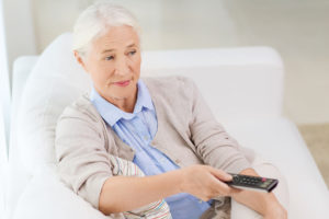 An elderly woman sits on a couch and points a remote toward a TV.