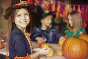 A young girl in a witch costume smiles as she holds a mini pumpkin. She wears no makeup.