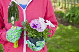 A person holds a shovel in one hand and a flower pot in the other. The person wears gardening gloves and stands outside.