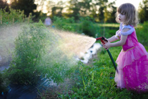 A little girl in a pink dress waters plants with a water spray gun.