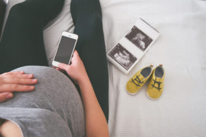 A pregnant woman sits on a bed and holds a smartphone in her hand. A picture of her baby and baby shoes sits on the bed next to her.