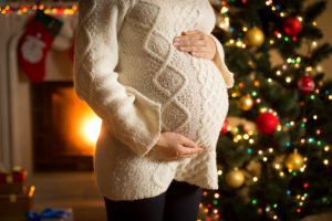 A pregnant woman stands next to a Christmas tree and holds her stomach.