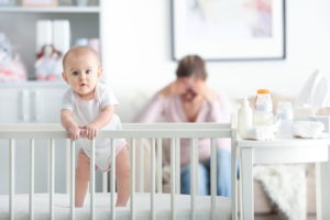 A baby sits in a crib. The mother sits near the crib and appears upset.