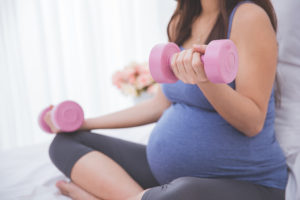 A pregnant woman crosses her legs as she sits.  She lifts light dumbbell weights.