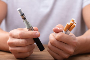 A person holds a e-cigarette in one hand and five cigarettes in the other.